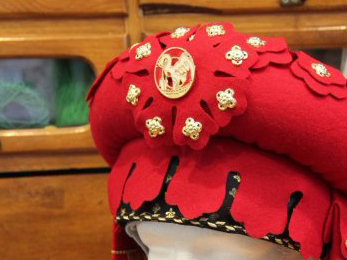 Part of a red hat made of a large stuffed roll, with a fancy cut fringe and gold decorations