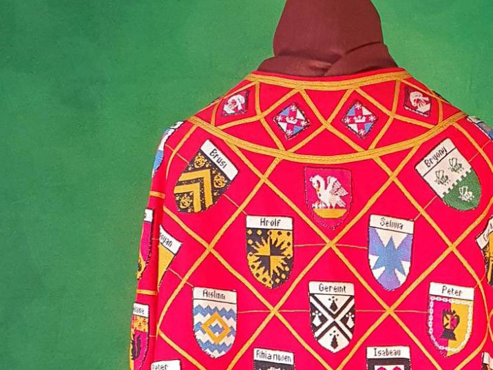 Top section of a cloak in red wool with many embroidered coats of arms in a grid formation