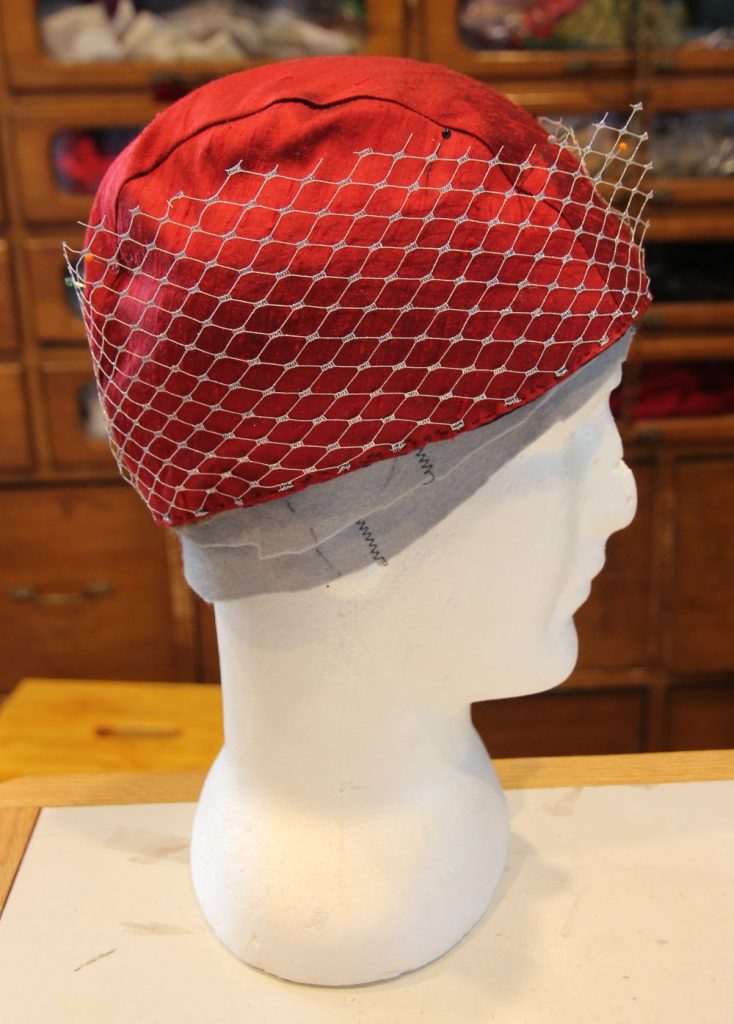 Headform wearing red silk cap with silver netting pinned to sides