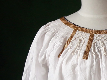 Shoulder of a mannequin wearing a gathered white linen underdress, with smocking and gold braid