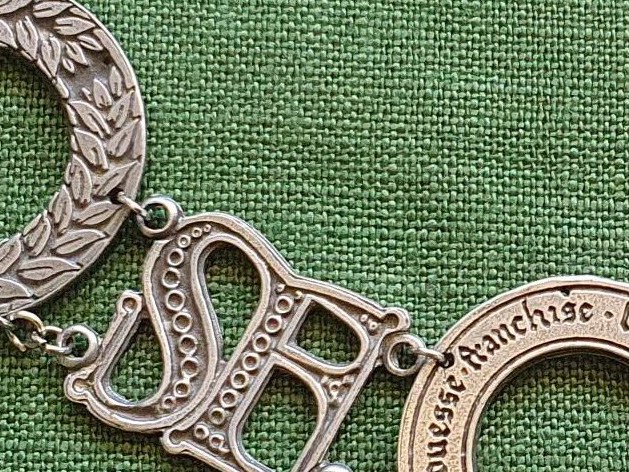 Part of a chain if medallions in pewter, showing a laurel wreath, SF and belt with the word Franchise