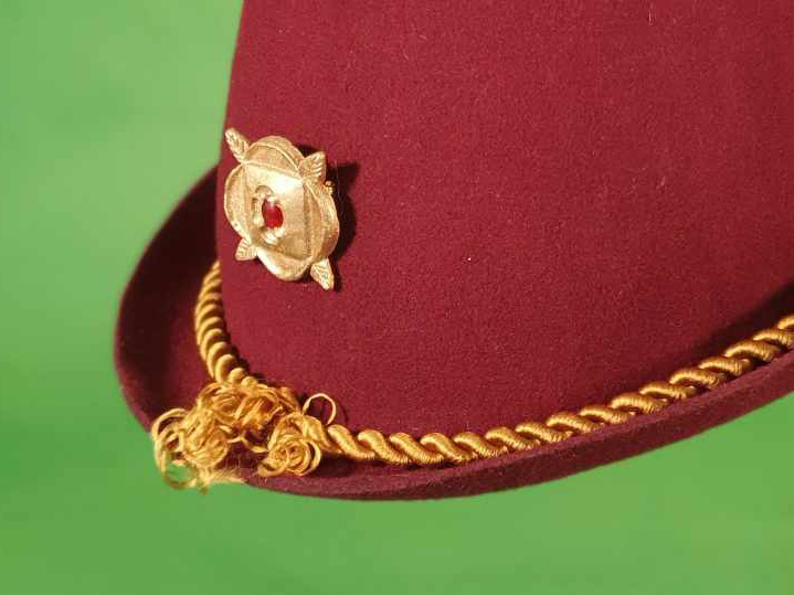 Front of a red felt hat with gold jewel brooch and gold cord hatband